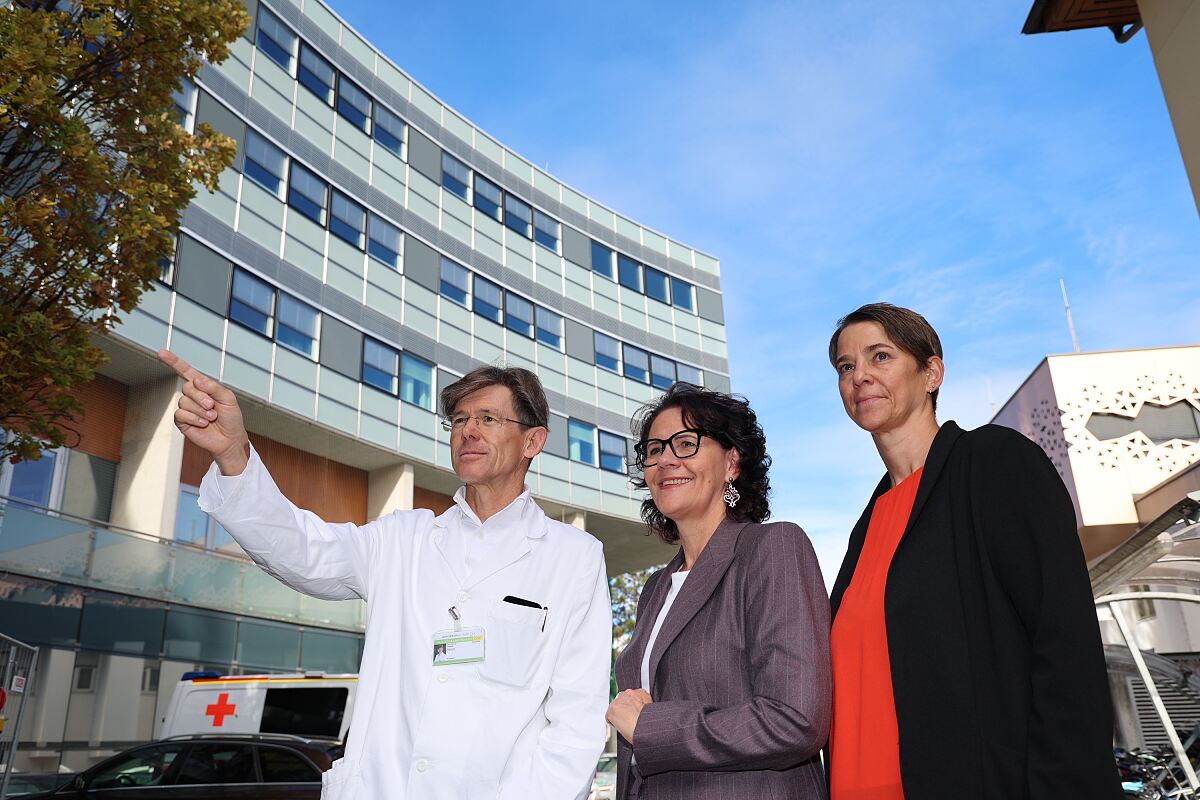 Arzt zeigt auf Krankenhausgebäude in Österreich mit zwei Kolleginnen
